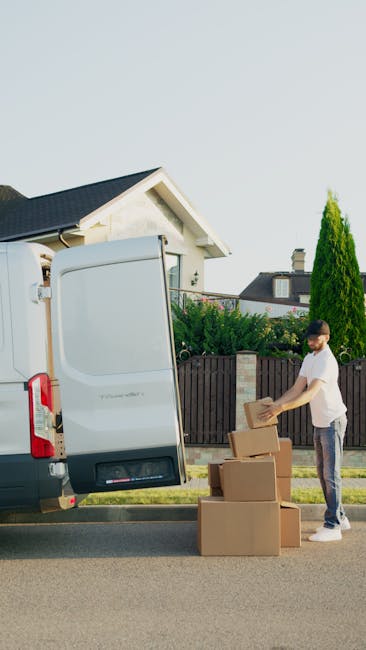 A man placing cardboard boxes onto the pavement beside the open rear doors of a white van, preparing for a house relocation. The scene is outdoors on a residential street, with a modern white house fe