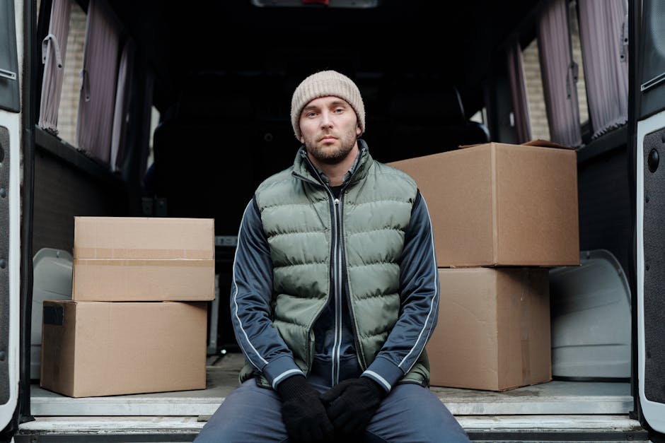 A man placing cardboard boxes onto the pavement beside the open rear doors of a white van, preparing for a house relocation. The scene is outdoors on a residential street, with a modern white house fe