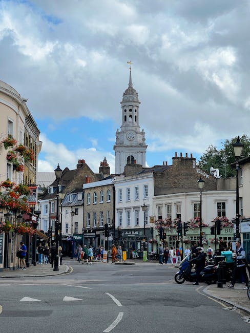 A street scene in Wimbledon Village with a mix of traditional and modern buildings, including shops and cafes, decorated with hanging flower baskets, with several pedestrians walking on the pavements and crossing the street. A motorbike and a cyclist are visible on the road, which has white pedestrian crossing markings. In the background, a tall, historic clock tower with a spire and weather vane rises above the surrounding rooftops, under a partly cloudy sky with patches of blue. Lantern-style street lighting is mounted on posts along the pavement. The scene captures an active, typical day in a busy residential and commercial area. Wimbledon Man and Van, a removals service, might be involved in local home relocations and furniture transport in this setting, supporting moving logistics and packing and moving activities.