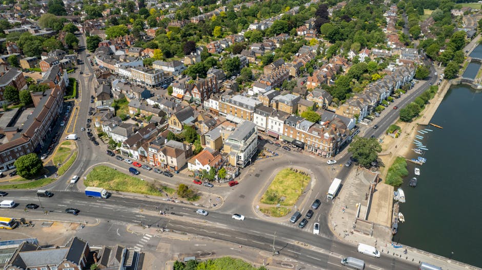 A street scene in Wimbledon Village with a mix of traditional and modern buildings, including shops and cafes, decorated with hanging flower baskets, with several pedestrians walking on the pavements and crossing the street. A motorbike and a cyclist are visible on the road, which has white pedestrian crossing markings. In the background, a tall, historic clock tower with a spire and weather vane rises above the surrounding rooftops, under a partly cloudy sky with patches of blue. Lantern-style street lighting is mounted on posts along the pavement. The scene captures an active, typical day in a busy residential and commercial area. Wimbledon Man and Van, a removals service, might be involved in local home relocations and furniture transport in this setting, supporting moving logistics and packing and moving activities.